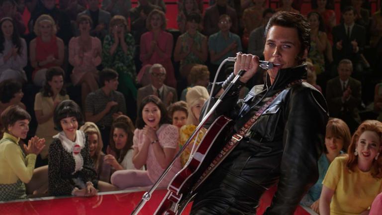 Austin Butler as Elvis Presley singing to a TV audience of adoring female fans in the movie ELVIS. He has a guitar strapped to him and is wearing a black leather suit.