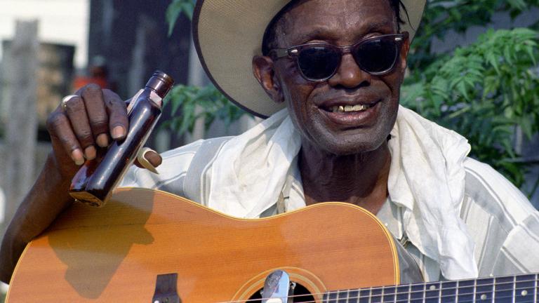 A middle aged African American man wearing a hat and sunglasses man holding a small bottle in one hand and a guitar.