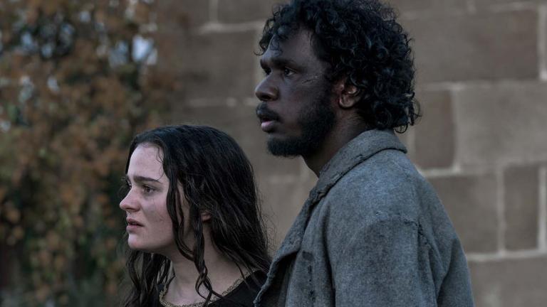 Aisling Franciosi and Baykali Ganambarr are standing outside in front of a wall in a scene from the film The Nightingale. They are looking pensively off camera