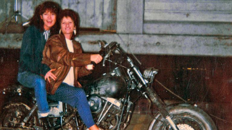 Two women sitting on a motorbike and smiling directly at the camera.