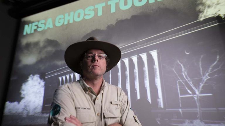 Tim the Yowie Man in the NFSA Theatrette standing before a slide of the NFSA during an evening Ghost Tour