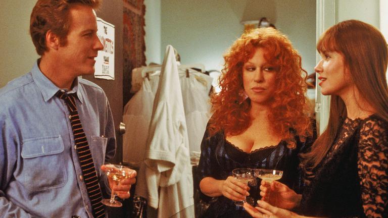 A man and two woman, roughly in their 30s, stand holding glasses of wine talking to each other in a scene from the film Beaches. One of the women looks unhappy.