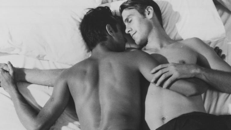 Two young men lying on a bed in an embrace. They are both shirtless and one is lying face down and the other facing up, with their faces turned toward eachother.
