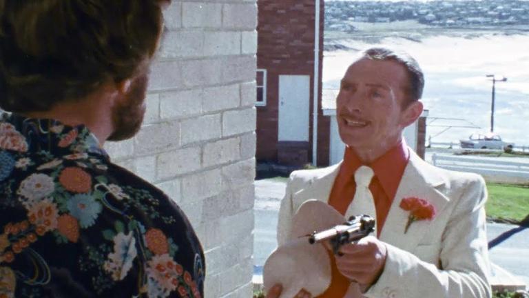 A man with his back to camera stands in a doorway to a building facing another man in a white suit who is pointing a gun at him.