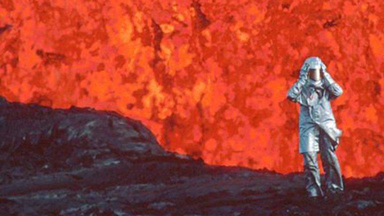 A person in a full body fire-protective suit walking near a volcano. You can see lava in the background.