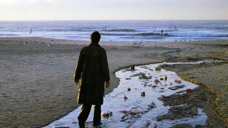 A man stands alone on an empty beach, wearing a long coat and with his back facing the camera. There is rubbish strewn on the beach in front of him.