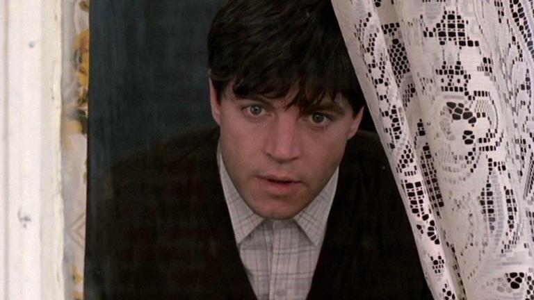 A young man peering out of a window through curtains.