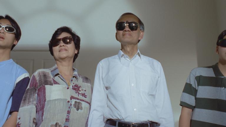 A family of four people standing side by side in a room looking outside. They are all wearing dark sunglasses.