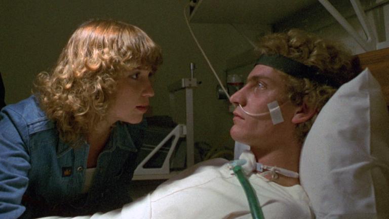 A man lies in a hospital bed, he is connected with tubes and machinery while a young woman stands next to the bed leaning over him.