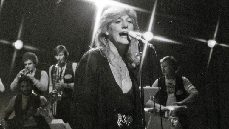 Singer Renee Geyer pictured from the waist up, singing into a microphone with band members in the background behind her.