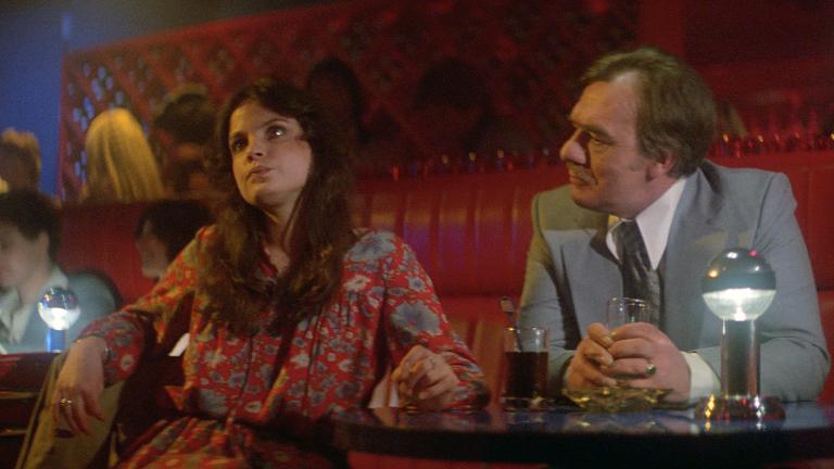 A young woman sits at what appears to be a table in a bar. She is looking unhappy with her head tilted away from an older man who is talking to her.