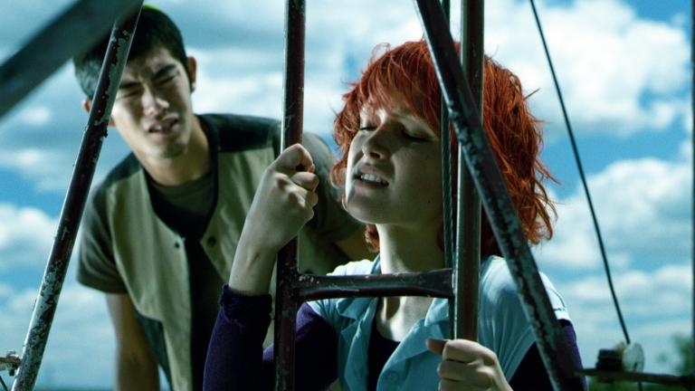 A confused-looking young man looks at a young woman who has her eyes closed and is holding on tightly to a metal ladder.