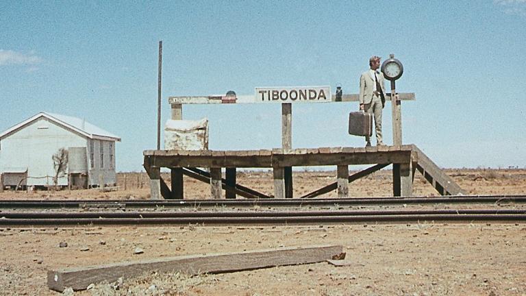 Wide shot of a man holding a suitcase is standing on a short platform at a train station in a remote outback area. The sign at the train station says Tiboonda.
