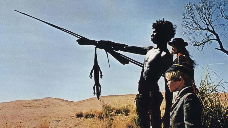 David Gulpilil, pointing a spear, stands in the desert with Jenny Agutter and Lucien John in private school uniforms in a scene from the film Walkabout