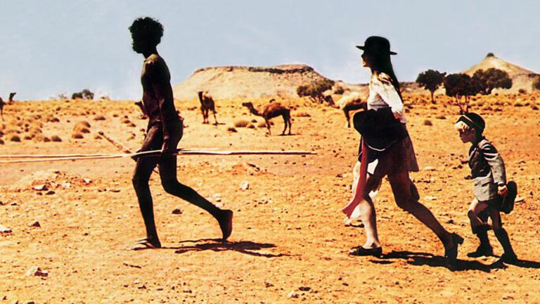 A young Aboriginal man holding a spear leading a teenage girl and young boy, walking in the outback in a scene from Walkabout.