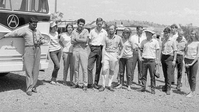 A group of 13 people standing side by side next to the front of a bus looking towards the camera.