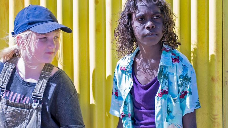 Two kids standing side by side against a yellow corrugated iron fence. One is white girl with straight blond hair in a ponytail, wearing denim overalls and a cap, and the other is a First Nations boy with curly dark hair wearing a purple singlet and blue Hawaiian shirt.