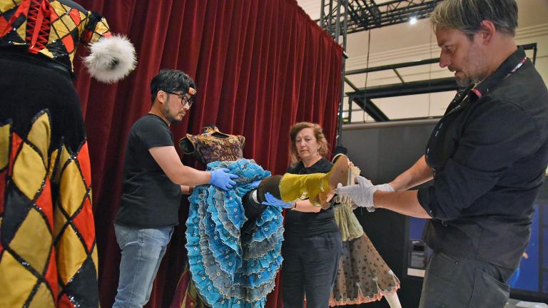 Three people on a stage putting together and posing a mannequin that is dressed in a costume from the film Moulin Rouge with a satin embroidered bodice and full layered skirt that has lace petticoats and long boots.