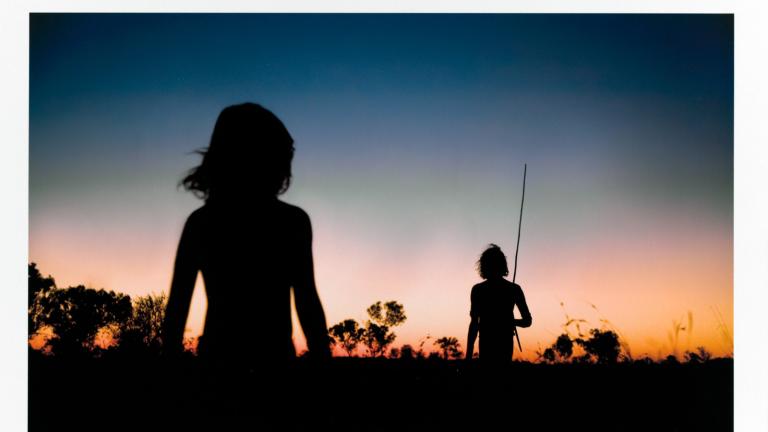 A boy and man in silhouette in the outback at dusk. The sky ranges from bright orange at the horizon to a deep blue above.  The photo has been signed in the bottom right corner by photographer Douglas Kirkland.