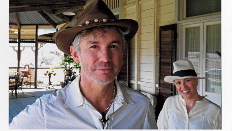 Head and shoulders shot of director Baz Luhrmann and production designer Catherine Martin. They are standing on a front porch of an old federation-style house, looking directly at the camera and smiling.  The photo has been signed in the bottom right corner by photographer Douglas Kirkland.