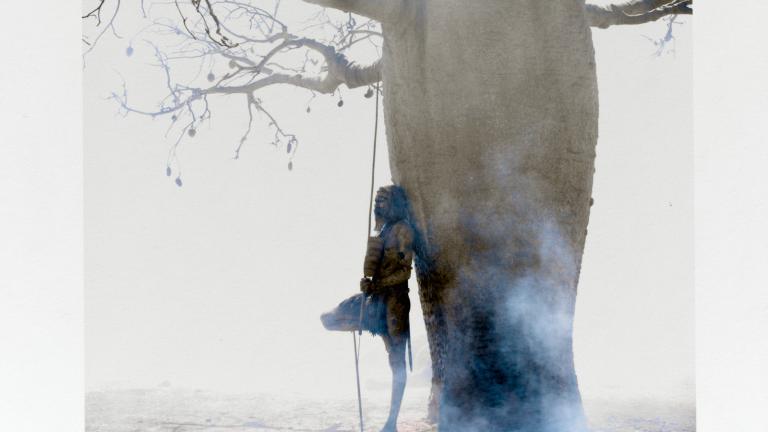 Actor David Gulpilil standing on one foot and leaning against a baobab tree in the outback during filming of Baz Luhrmann's film Australia.  The photo has been signed in the bottom right corner by photographer Douglas Kirkland.