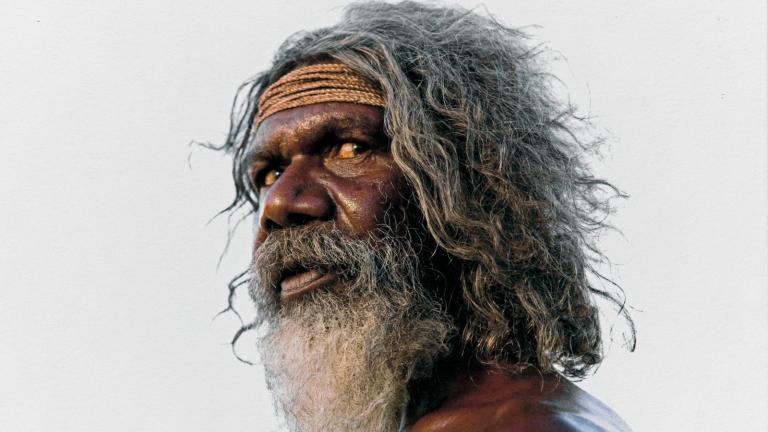 Head and shoulders shot of actor David Gulpilil standing side-on and looking to his left toward the camera. He has long hair and a beard, and has a large vertical scar on his shoulder. The photo has been signed in the bottom right corner by photographer Douglas Kirkland.