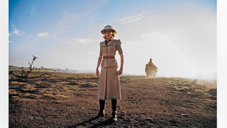 Actor Nicole Kidman wearing a safari-style outfit with a long skirt, belted shirt and knee-high boots and a hat. She is standing in a dusty outback setting during the filming of Baz Luhrmann's film Australia.  The photo has been signed in the bottom right corner by photographer Douglas Kirkland.