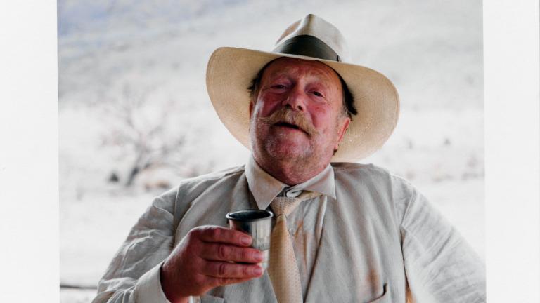 Actor Jack Thompson standing leaning against a railing. He is wearing a hat, vest and tie and holding a cup in one hand.  The photo has been signed in the bottom right corner by photographer Douglas Kirkland.
