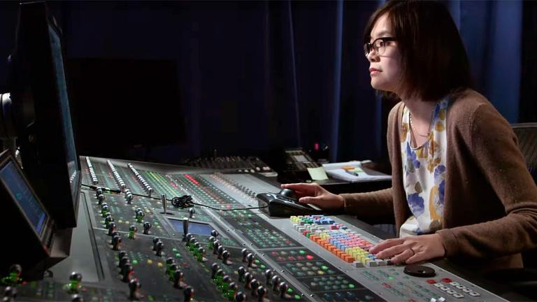 A woman sitting in a studio at a sound desk looking at a screen, there are a lot of buttons and dials in front of her. 
