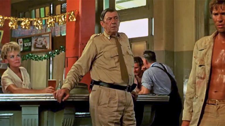 A middle-aged policeman leaning against a bar in a country pub looking at a young man covered in sweat and blood. Behind the bar a woman is serving a beer. The image is a scene from the film Wake in Fright. 