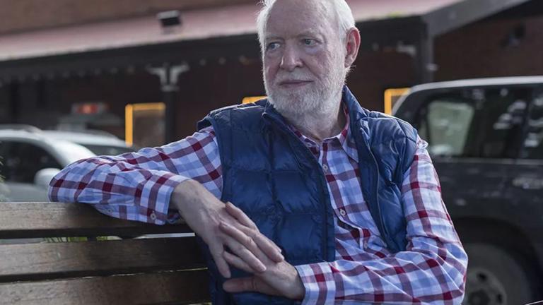 Film critic and author David Stratton seated on a bench outdoors. He is wearing a check shirt and vest and looking off to his right. 