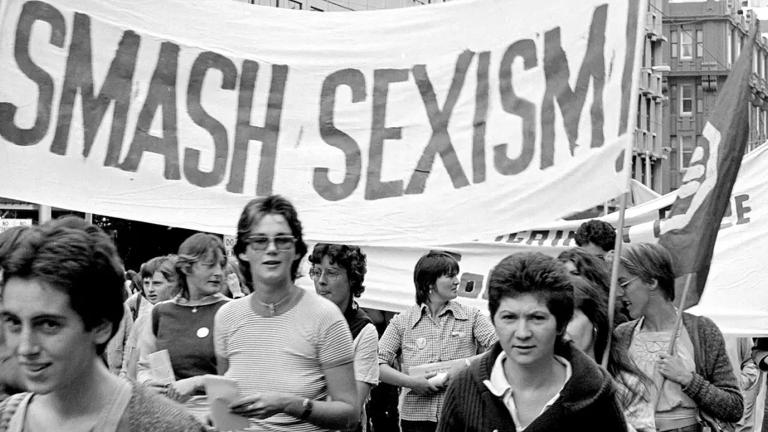 Women marching in the street with a big banner that reads SMASH SEXISM.