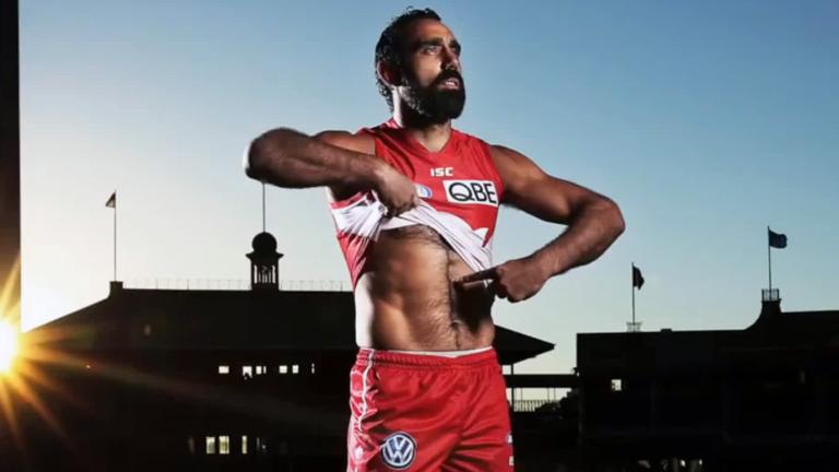 AFL football player Adam Goodes in the middle of a sports ground. He is wearing his Sydney Swans red and white uniform and lifting up the front of his shirt to point to the colour of his skin. 