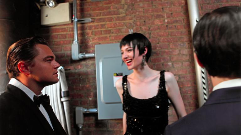 Actor Leonardo DiCaprio wearing a tuxedo standing side-on facing right of screen, Actor Elizabeth Debicki in a black sequinned dress is looking at him smiling and another man with his back to the camera is standing right of screen.