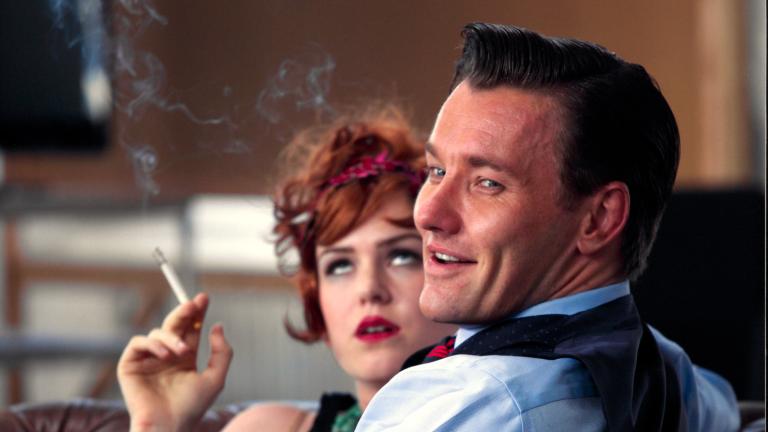 Actors Joel Edgerton and Isla Fisher dressed in 1920s costume, seated on a couch.