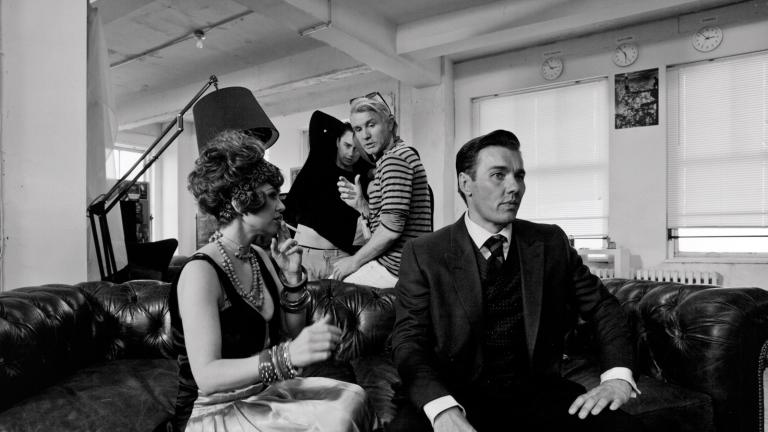 Actors Isla Fisher and Joel Edgerton wearing 1920s costumes and seated on a big leather couch facing towards something outside the frame. Director Baz Luhrmann is behind them looking towards them.