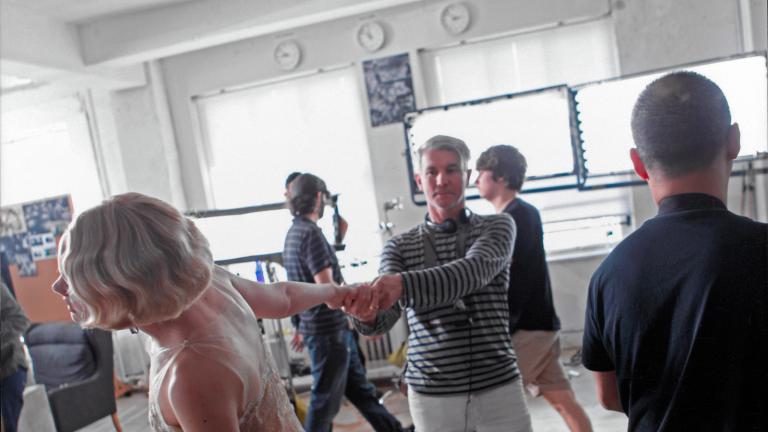 Actor Carey Mulligan facing away from the camera wearing a 1920s-style dress. Director Baz Luhrmann is holding her outstretched hand and they are in a large room surrounded by film production crew members.