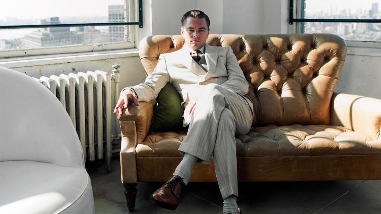 Actor Leonardo DiCaprio in a behind the scenes still from the film The Great Gatsby. He is wearing a white suit and is seated on a tan-coloured couch in the corner of a room. He's looking directly at the camera. Out of the windows behind him you can see a city skyline. 