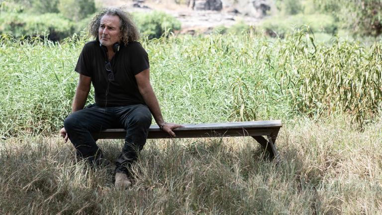 Director Stephen Maxwell Johnson sitting on a wooden bench in a grassy area on the set of his film High Ground. 