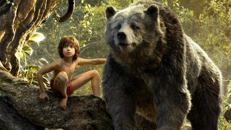 A young boy sitting on at the foot of a tree next to a huge grizzly bear in a scene from The Jungle Book film.