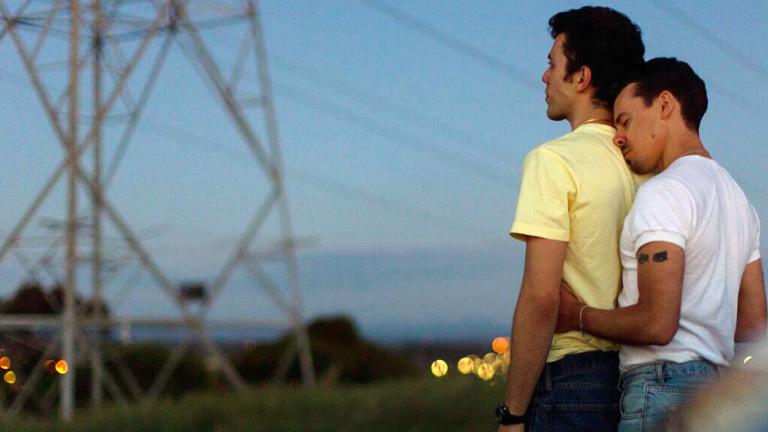 A young man holds another young man around the waist from behind. They are standing outside with a view of an electrical tower