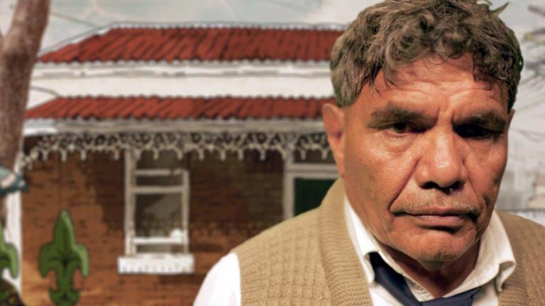 Head and shoulders shot of a solemn-looking middle-aged First Nations man looking directly at camera. There is a small brick house in the background behind him.