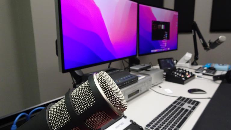 A large radio microphone in the foreground, in the background are some computer monitors and keyboards on a desk.
