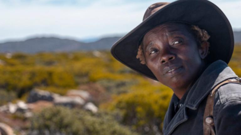 Head and shoulders shot of a Nigerian woman wearing a coat and hat with an outback landscape of hills and mountains behind her.