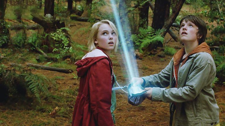 Two young teens are in a forest looking up towards the sky at a bright light beaming down in front of them.