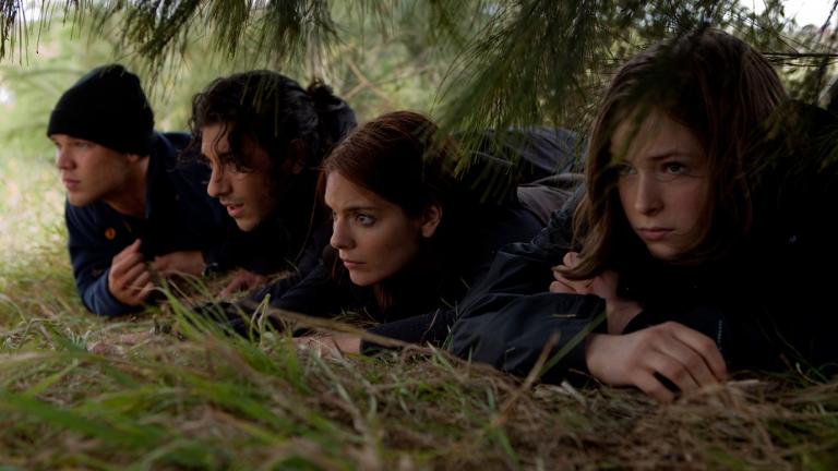Four teenagers lying on the ground, trying to hide themselves under trees and scrub as they watch over something happening off screen.