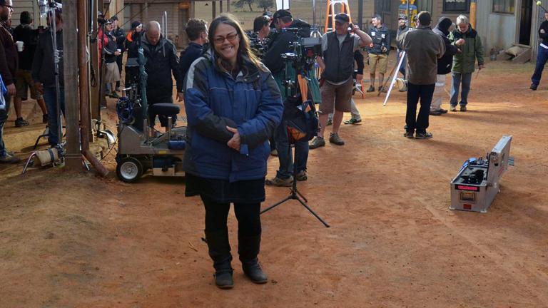 Director Jocelyn Moorhouse on the set of a film. She is looking at the camera while a large film crew are at work behind her. 