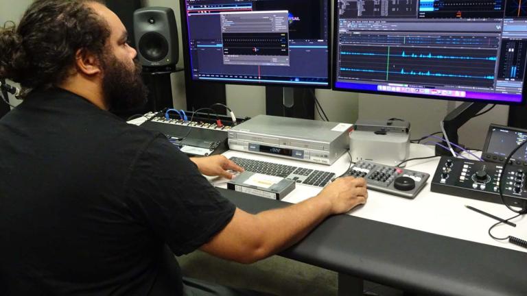A First Nations man seated at a desk in a studio covered in computers and recording equipment