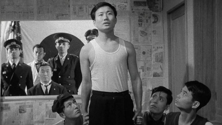 A young Japanese man standing in a room surrounded by three other men who are holding onto his hands. In the back of the room are a group of policemen.