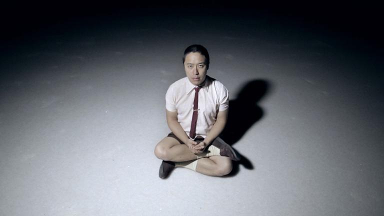 An Asian man sitting cross-legged on a floor in a dark room with a spotlight on him. He is wearing a short-sleeve shirt with a tie and shorts and he is looking up at the camera.
